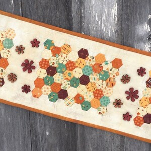 May include: A beige table runner with a brown border features a patchwork design of hexagons in shades of orange, green, brown, and cream. The runner is decorated with wooden flower-shaped buttons in shades of brown and red.