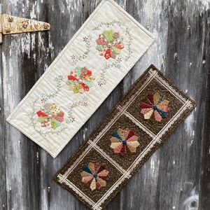 May include: Two table runners with a floral design. One runner is white with green vines and three multi-colored flowers. The other runner is brown with white lace trim and three multi-colored flowers.