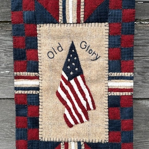 May include: A rectangular wall hanging featuring a central tan panel with a small American flag and the text Old Glory embroidered in black. The border consists of a red and navy blue checkered pattern with tan and white striped accents.