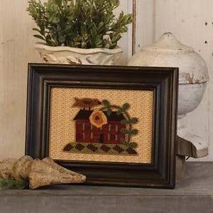 May include: A framed, rustic artwork featuring a red house with a sunflower and green foliage. The artwork is set against a tan background and framed in a dark brown wooden frame. Decorative elements include a white pot with greenery and a white, aged decorative piece.