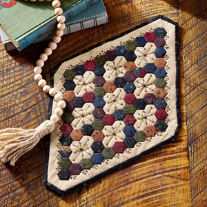 May include: A handcrafted, quilted table runner with a patchwork design of hexagonal fabric pieces in various colors, including burgundy, navy, and olive green. The runner features embroidered floral accents and a cream-colored border with a dark blue trim. A wooden bead garland with a tassel is also present.