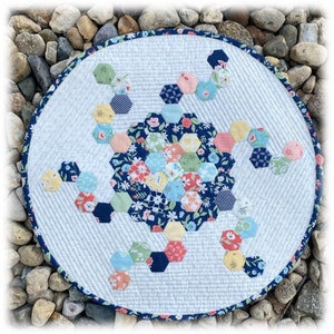 May include: A white round quilted table runner with a blue border. The center of the runner features a hexagonal design with a variety of floral fabrics in shades of blue, pink, yellow, and green.