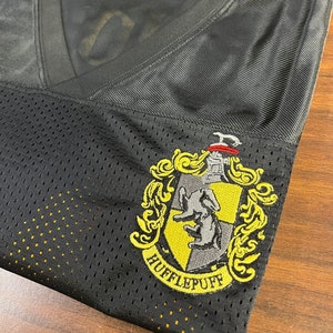 Pu&ograve; includere: Maglia in mesh nera con uno stemma di Hufflepuff ricamato in giallo e grigio. Lo stemma presenta un tasso e le parole "Hufflepuff" in un arco giallo.