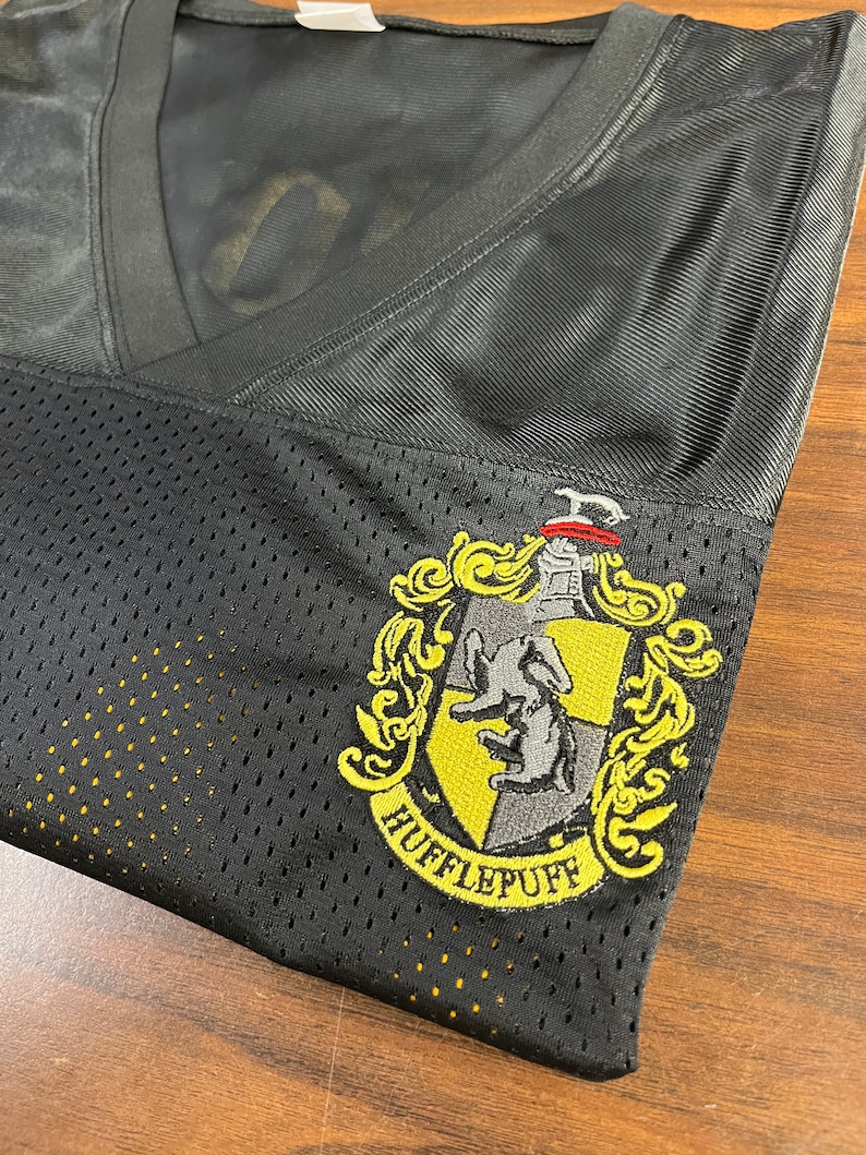 Pu&ograve; includere: Maglia in mesh nera con uno stemma di Hufflepuff ricamato in giallo e grigio. Lo stemma presenta un tasso e le parole "Hufflepuff" in un arco giallo.