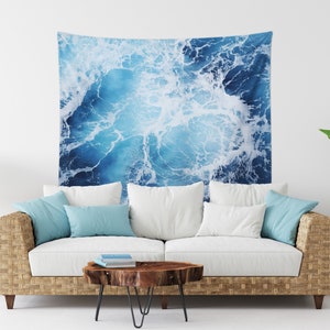May include: A blue and white tapestry featuring a close-up of ocean waves crashing. The tapestry is hanging on a white wall above a white couch with blue throw pillows.