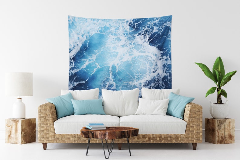 May include: A blue and white tapestry featuring a close-up of ocean waves crashing. The tapestry is hanging on a white wall above a white couch with blue throw pillows.