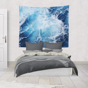 May include: A gray bed with white bedding and a blue and white tapestry hanging on the wall behind it. The tapestry features a close-up of ocean waves.