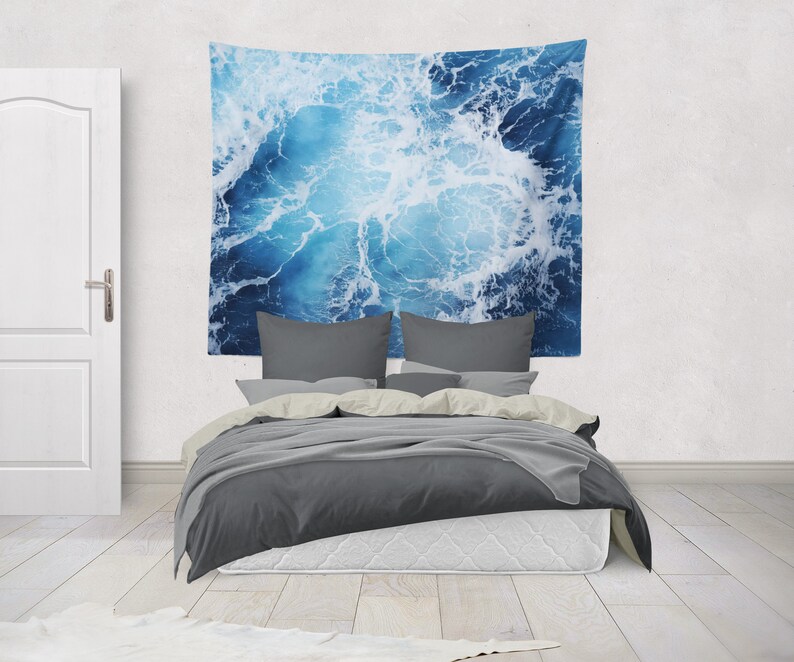 May include: A gray bed with white bedding and a blue and white tapestry hanging on the wall behind it. The tapestry features a close-up of ocean waves.