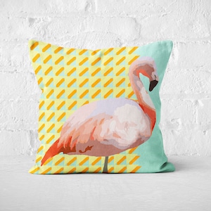 Puede incluir: Una almohada de flamenco rosa con un patrón geométrico amarillo y azul. La almohada está sobre un fondo de ladrillo blanco.