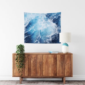 May include: A blue and white tapestry featuring a close-up of ocean waves with white foam. The tapestry is hanging on a white wall above a wooden cabinet with a plant and a lamp.