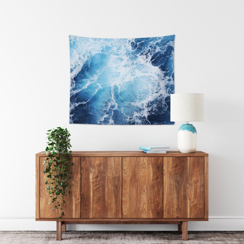 May include: A blue and white tapestry featuring a close-up of ocean waves with white foam. The tapestry is hanging on a white wall above a wooden cabinet with a plant and a lamp.