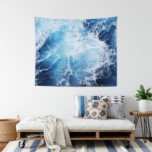 May include: A blue and white tapestry featuring a close-up of ocean waves with white foam. The tapestry is hanging on a white wall above a white couch with blue and white throw pillows.