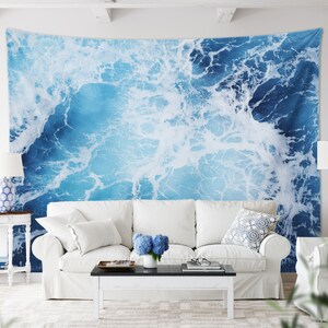 May include: A white couch with white throw pillows and a blue and white patterned throw pillow sits in front of a wall tapestry depicting a blue ocean with white foam.