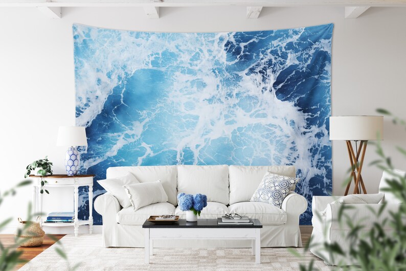 May include: A white couch with white throw pillows and a blue and white patterned throw pillow sits in front of a wall tapestry depicting a blue ocean with white foam.