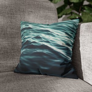 May include: Square cushion featuring a digitally printed ocean wave design in teal and navy blue hues. The cushion is placed on a grey upholstered sofa, bringing a coastal theme to the living room.