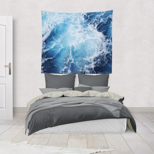 May include: A gray bed with a white headboard and a blue and white tapestry hanging above it. The tapestry features a close-up of ocean waves.