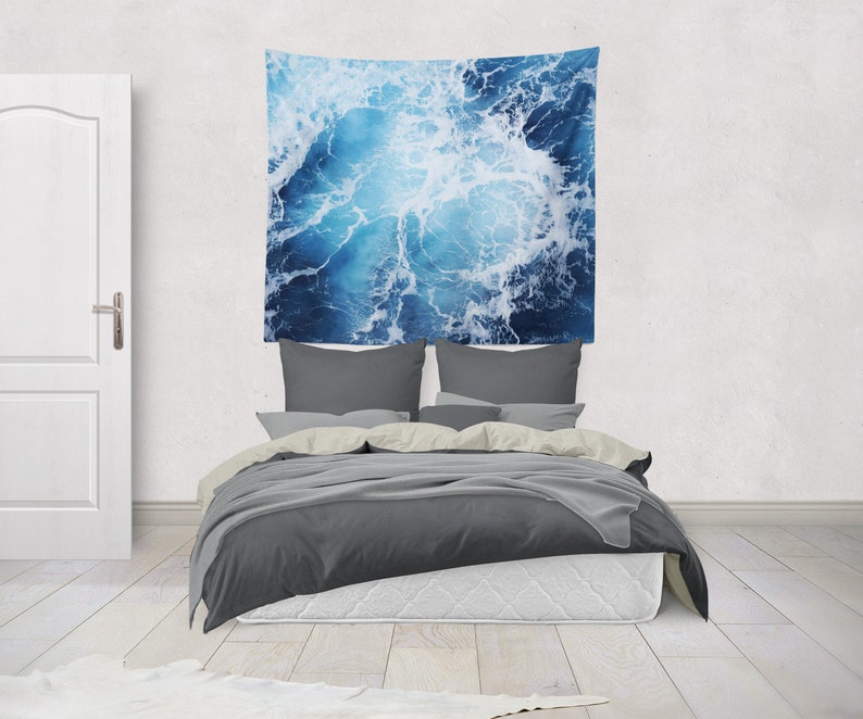 May include: A gray bed with a white headboard and a blue and white tapestry hanging above it. The tapestry features a close-up of ocean waves.