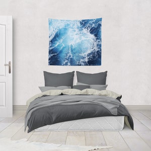 May include: A gray bed with white bedding and two gray pillows. A blue and white tapestry with a wave pattern hangs on the wall above the bed.