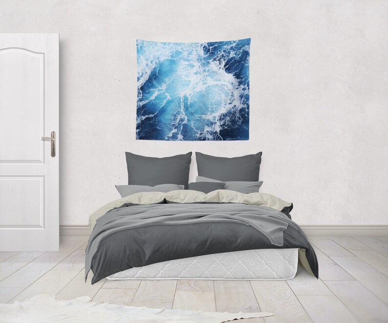 May include: A gray bed with white bedding and two gray pillows. A blue and white tapestry with a wave pattern hangs on the wall above the bed.