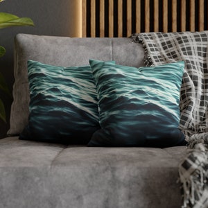 May include: Two square cushions featuring a teal and dark blue water wave design. They rest on a grey sofa, with a grey headrest and a black and white checked blanket.