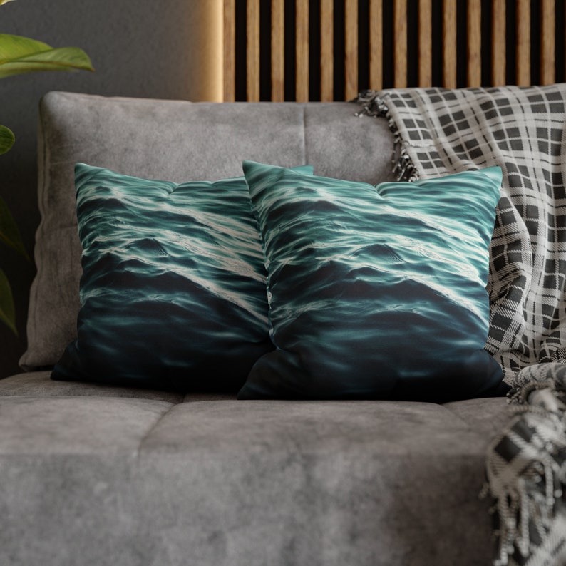 May include: Two square cushions featuring a teal and dark blue water wave design. They rest on a grey sofa, with a grey headrest and a black and white checked blanket.
