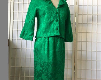 May include: A green vintage skirt suit with a floral pattern. The jacket has a ruffled collar and three buttons. The skirt is knee-length.