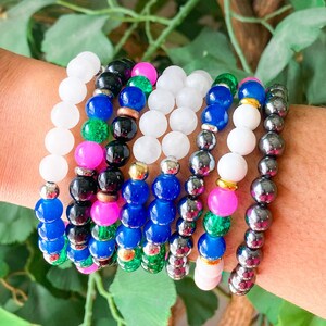 May include: A collection of beaded bracelets in various colors, including white, black, blue, pink, and green. The bracelets are strung together with silver-colored beads. The background features green foliage.