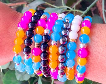 Può includere: Una collezione di braccialetti di perline in vari colori, tra cui rosa, blu, giallo, arancione, nero e bianco. I braccialetti sono indossati insieme su un polso, mostrando un accessorio vivace.