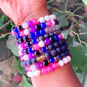 May include: A collection of beaded bracelets in various colors, including pink, blue, white, black, and purple. Some bracelets feature gold-colored accents. The bracelets are stacked on a wrist, showcasing the variety of designs and colors.