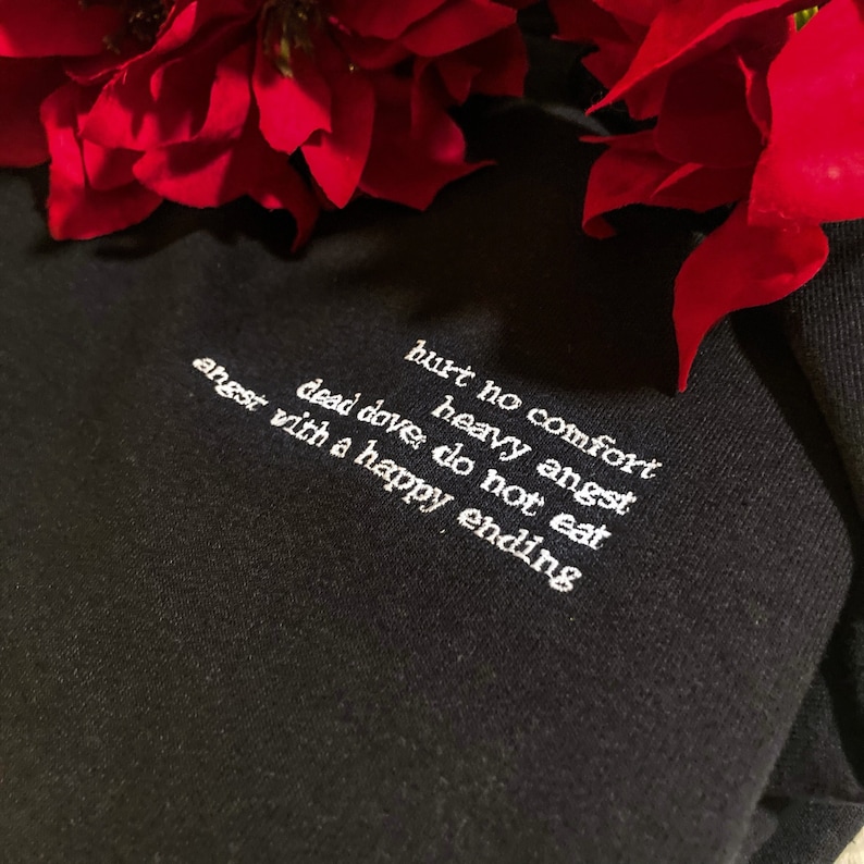 May include: Black sweatshirt with white embroidered text: "hurt no comfort, heavy angst, dead dove do not eat, angst with a happy ending." Red flowers are in the background.