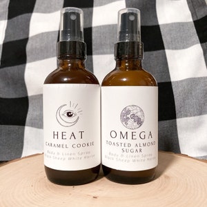 May include: Two amber glass bottles with black spray tops and white labels. The labels feature a crescent moon with an eye inside and the text "HEAT CARAMEL COOKIE Body & Linen Spray Black Sheep White Horse" on the left bottle. The right bottle has a crescent moon with a stylized flower inside and the text "OMEGA TOASTED ALMOND SUGAR Body & Linen Spray Black Sheep White Horse".