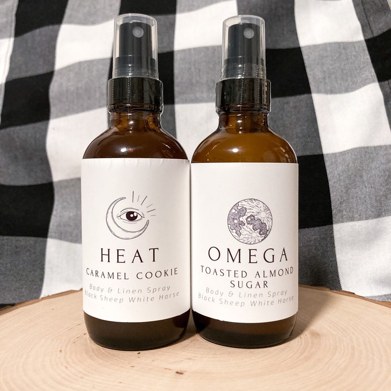 May include: Two amber glass bottles with black spray tops and white labels. The labels feature a crescent moon with an eye inside and the text "HEAT CARAMEL COOKIE Body & Linen Spray Black Sheep White Horse" on the left bottle. The right bottle has a crescent moon with a stylized flower inside and the text "OMEGA TOASTED ALMOND SUGAR Body & Linen Spray Black Sheep White Horse".