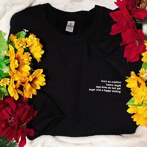 May include: Black sweatshirt with white embroidered text: "hurt no comfort, heavy angst, dead dove do not eat, angst with a happy ending." The sweatshirt is surrounded by yellow and red flowers.
