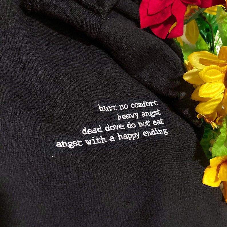 May include: Black hoodie with white embroidered text. The text reads "hurt no comfort, heavy angst, dead dove do not eat, angst with a happy ending." The hoodie is made of a soft, dark fabric.
