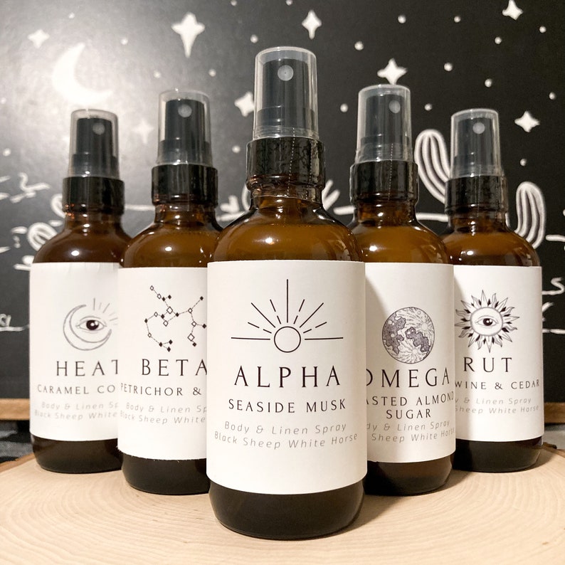 May include: Five amber glass bottles with black spray tops and white labels. Each bottle has a different design and text. The text on the labels reads: "HEAT CARAMEL CO", "BETA PETRICHOR &", "ALPHA SEASIDE MUSK", "OMEGA TOASTED ALMOND SUGAR", and "RUT WINE & CEDAR".