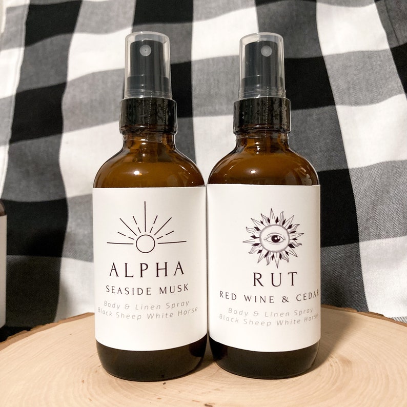 May include: Two amber glass bottles with white labels and black spray tops. The labels feature a sun and eye graphic with the text "ALPHA SEASIDE MUSK Body & Linen Spray Black Sheep White Horse" and "RUT RED WINE & CEDAR Body & Linen Spray Black Sheep White Horse".