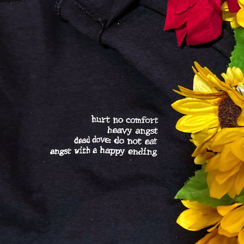 May include: Black sweatshirt with white embroidered text: "hurt no comfort, heavy angst, dead dove do not eat, angst with a happy ending." The sweatshirt is next to yellow sunflowers and red flowers.