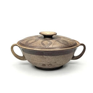 May include: A ceramic soup tureen with a lid. The tureen is a speckled tan color with a dark brown band around the middle. The lid has a repeating pattern and a small knob. The tureen has two handles.