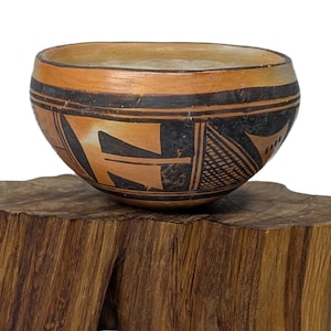 May include: A handcrafted, round ceramic bowl with a tan and black geometric design, resting on a rustic, wooden base. The bowl's exterior features a band of black geometric patterns and a tan background. The wooden base has a natural, rough-hewn appearance.