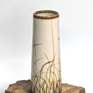May include: A cylindrical ceramic vase with a cream-colored glaze and a hand-painted brown grass motif. The rim features a dark brown drip glaze detail. The vase has a smooth, tapered body and a flat base, showcasing a naturalistic botanical design.
