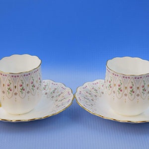 May include: Two white teacups and saucers with gold handles and rims. The cups and saucers have a scalloped edge and are decorated with pink floral and gold accents. The background is a gradient blue.