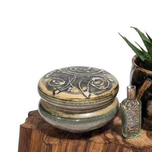 May include: A ceramic jar with a lid, a small glass bottle, and a potted succulent plant sit on a wooden surface. The jar has a gray and beige design. The bottle is clear. The plant pot is brown.