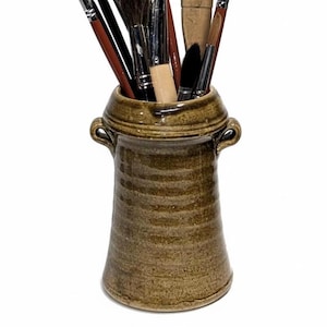 May include: A collection of paint brushes of various sizes and shapes, displayed in a brown ceramic jar. The brushes have wooden handles and natural bristles. The jar has a textured surface and two small handles.