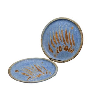 May include: Two round ceramic plates featuring a speckled light blue glaze with a series of vertical, elongated brown brushstroke patterns. The plates have a visible, textured rim and a consistent color palette across both pieces.