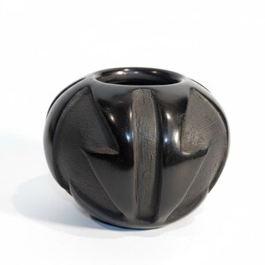 May include: A black, spherical vase with a textured, carved design. The vase has a wide opening and a glossy finish. The carved pattern features vertical and angled lines, creating a geometric effect. The vase is likely made of ceramic or stone.