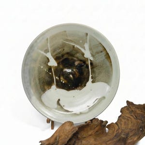 May include: A ceramic plate with a unique design. The plate features a dark brown center with white and light brown accents. The rim is a light gray color. The plate is displayed on a wooden stand.