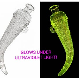 May include: Clear glass bottle with a curved, horn-like shape and a handle. The bottle has a textured, patterned surface. The text "GLOWS UNDER ULTRAVIOLET LIGHT!" is visible. The bottle glows bright green under UV light.