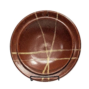 May include: A round, brown ceramic plate with a slightly raised rim. The plate features a series of intersecting, light-colored lines across its surface. The plate is displayed on a black metal stand, showcasing its design and texture.