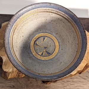 May include: A ceramic bowl with a speckled gray interior and a blue rim. The bowl features gold-colored concentric circles and a central design. The bowl is displayed on a piece of wood.