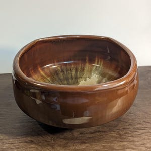 May include: A brown ceramic bowl with a glossy finish. The interior features a blend of green and beige hues, creating a unique pattern. The bowl has a rounded shape with a slightly squared rim, suitable for serving or display.